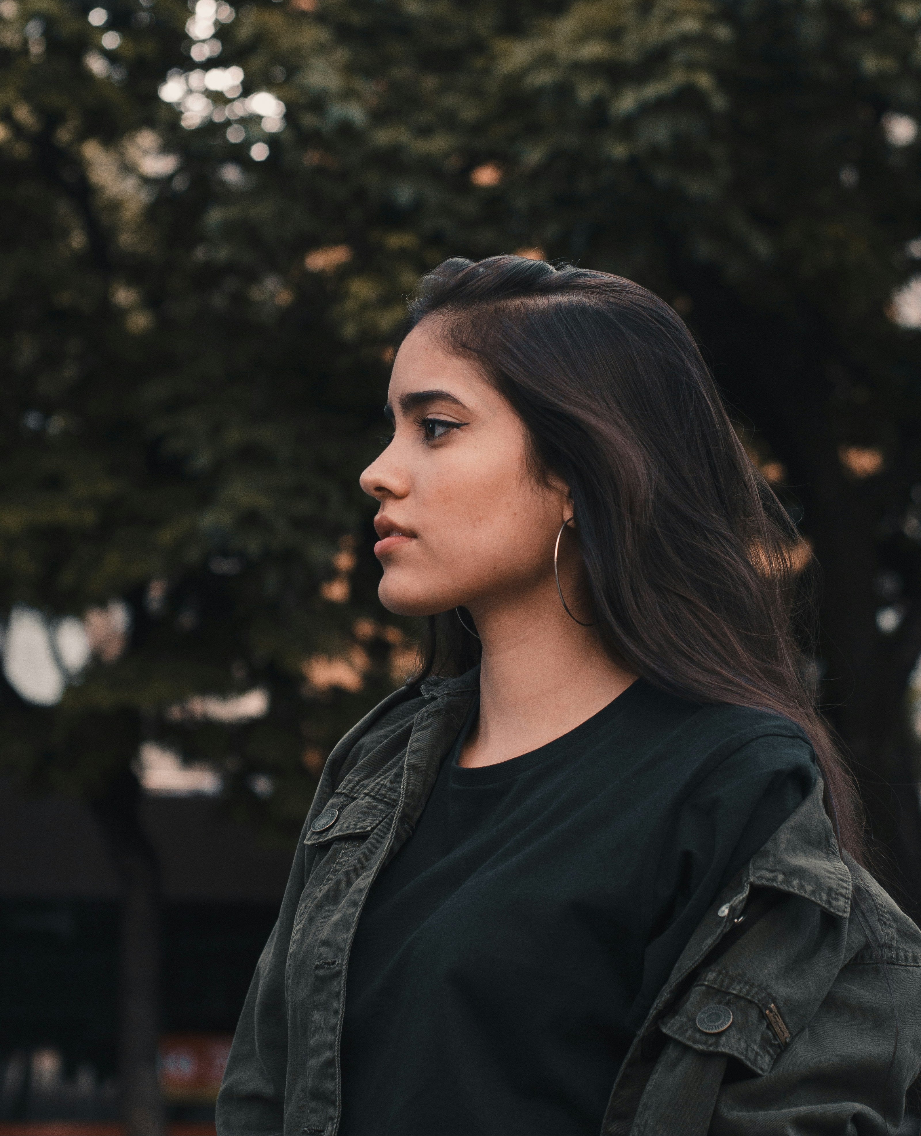 Woman with long dark hair, courtsey of kaers black seed hair oil, wearing a green jacket and black top, standing against a blurred natural background.
