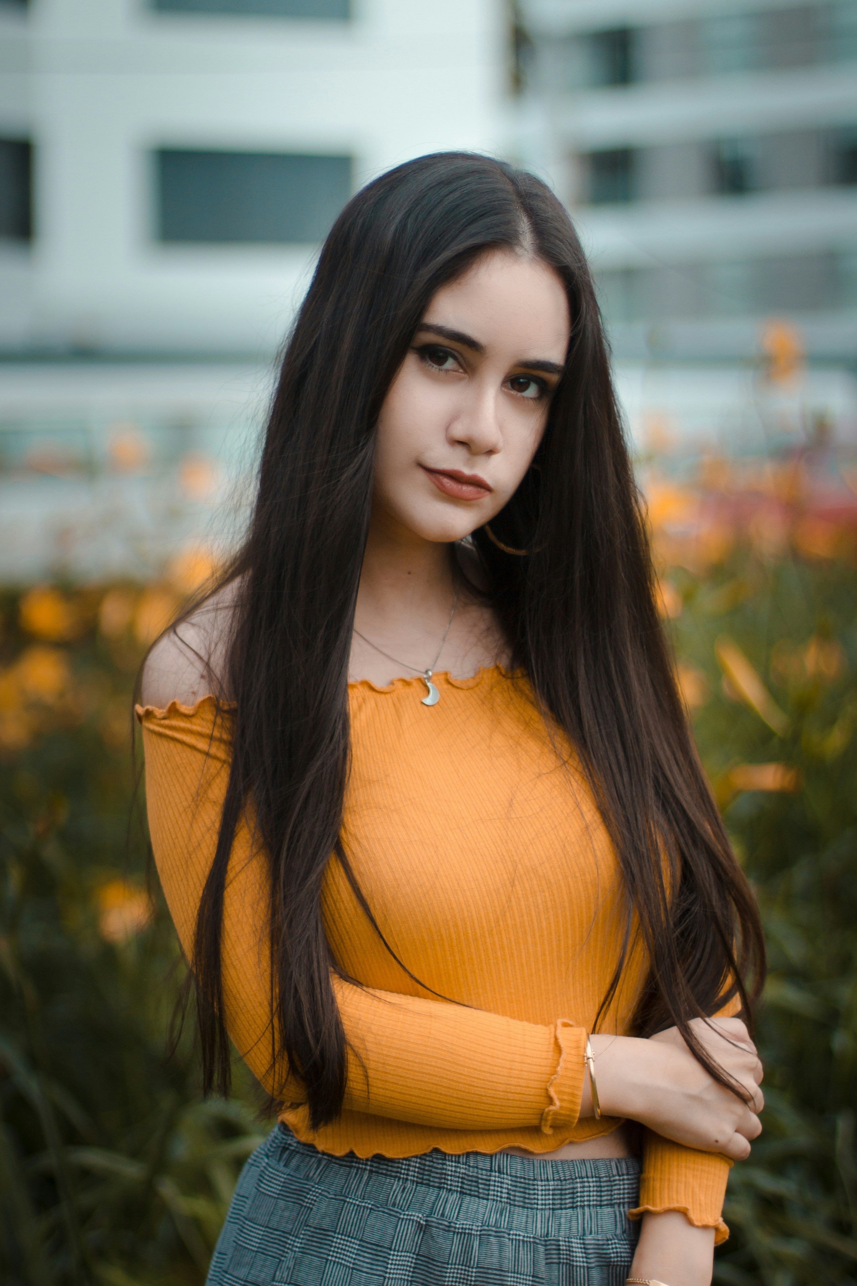 Woman in an orange top and blue plaid skirt standing in a blurred outdoor setting, with beautiful hair achieved by kaers haircare