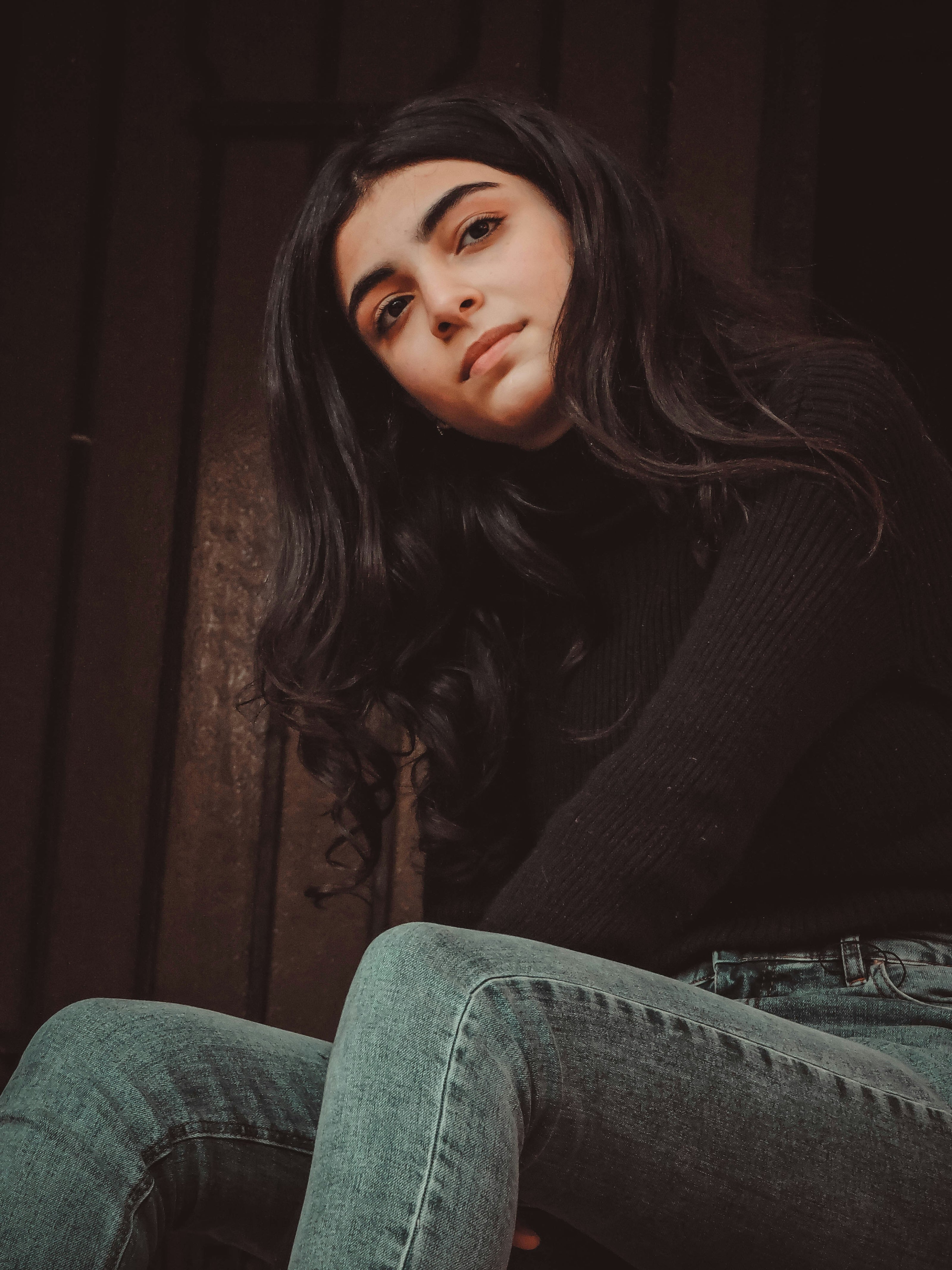 Person with kaers long dark hair wearing a black sweater and jeans against a dark background
