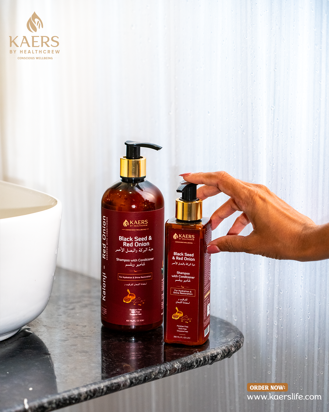 Two bottles of KAERS Black Seed & Red Onion hair care products on a marble surface with a hand reaching for one bottle.